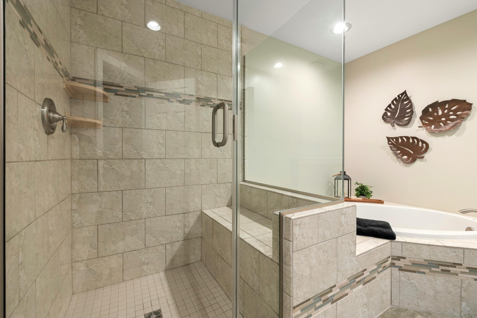 A stylish beige tiled bathroom featuring a glass shower and bathtub with decorative brown leaf art on the wall.